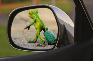 Green ceramic frog moving away pulling suitcase, as seen through car side mirror