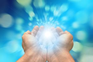 White hands receiving light from above, with a background of dark blue and light blue blurry spheres