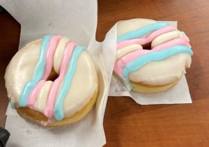 Brown table with a white napkin, two white doughnuts with white frosting and blue, pink and white icing in the shape of the trans flag