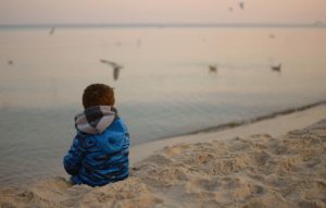 child in a blue jacket sitting on a sandy beach looking at seagulls, fuzzy horizon