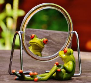Green ceramic frog with red eyes and toes looking at itself in a round mirror