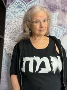 Silver and purple haired femme wearing a black tank top with the word EMET on it in white Hebrew letters, in front of a purple and  blue mandala background