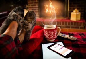 Comfy scene in various shades of red, with a fire in the background, a person's feet in pyjamas and slippers, a warm beverage, a phone and a remote