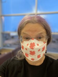 Silver and purple haired femme in front of a window with blue sky, wearing a white and red maple leaf KN95 respirator