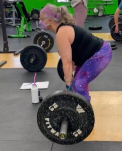 Silver haired femme wearing a black top and purple leggings, headband and mask, doing a deadlift