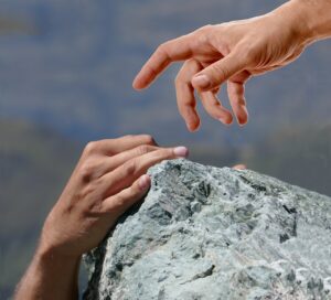 Grey and blue rock climbing scene, with one hand clinging to the edge of a rock and the second hand reaching down help.