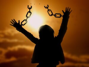 Orangey background with blurry sun, silhouette of a person with long hair and long sleeves, arms up, chains on their wrists trailing up into nothing