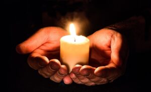 Black background, two hands holding a lit votive candle