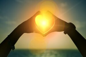 Beach scene with a setting sun, with a pair of hands making a heart around the sun.